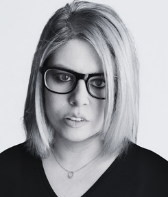 Maria looking straight into the camera, wearing thick black glassess and a black top